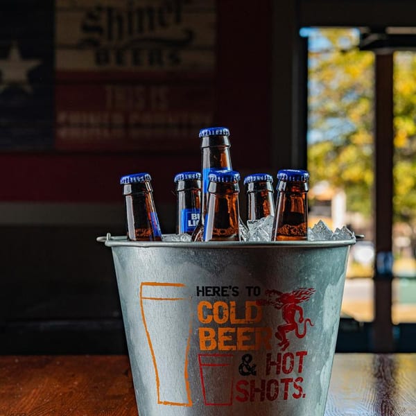 A metal bucket of bottled beers on ice, with "Here's to Cold Beer & Hot Shots" text.