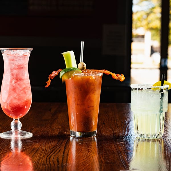 Three cocktails on a dark wooden bar: a red drink, a Bloody Mary with bacon, and a margarita.