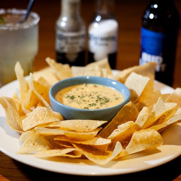 A plate of tortilla chips with a bowl of queso dip, a margarita, and beer bottles.