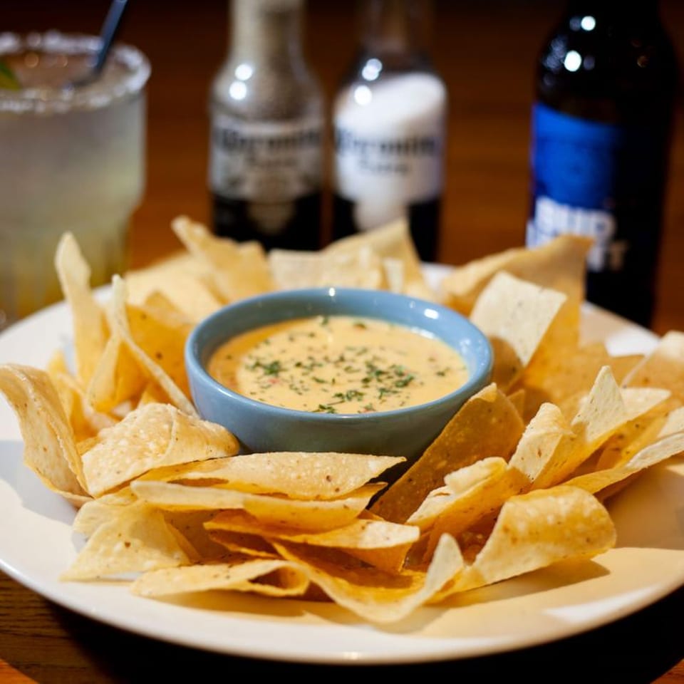 A plate of tortilla chips with a bowl of queso dip, a margarita, and beer bottles.