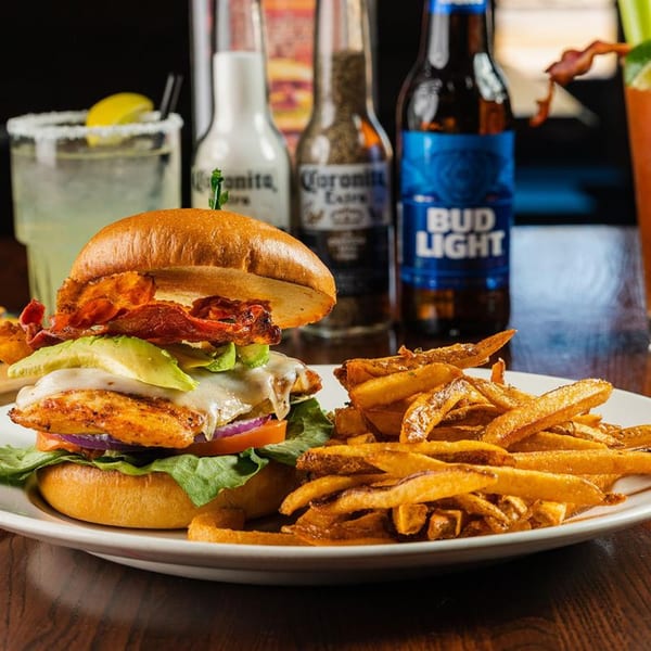 Chicken sandwich with bacon, avocado, and cheese, served with fries, margarita, and beer bottles.
